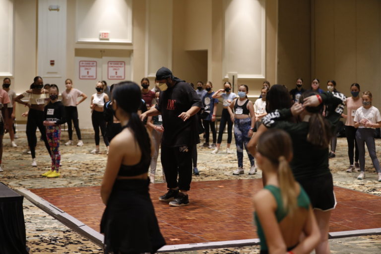 Justin Streeter, a Black man wearing all an all-black outfit stands in a hotel ballroom, speaking as dozens of masks dancers stand around him, listening.