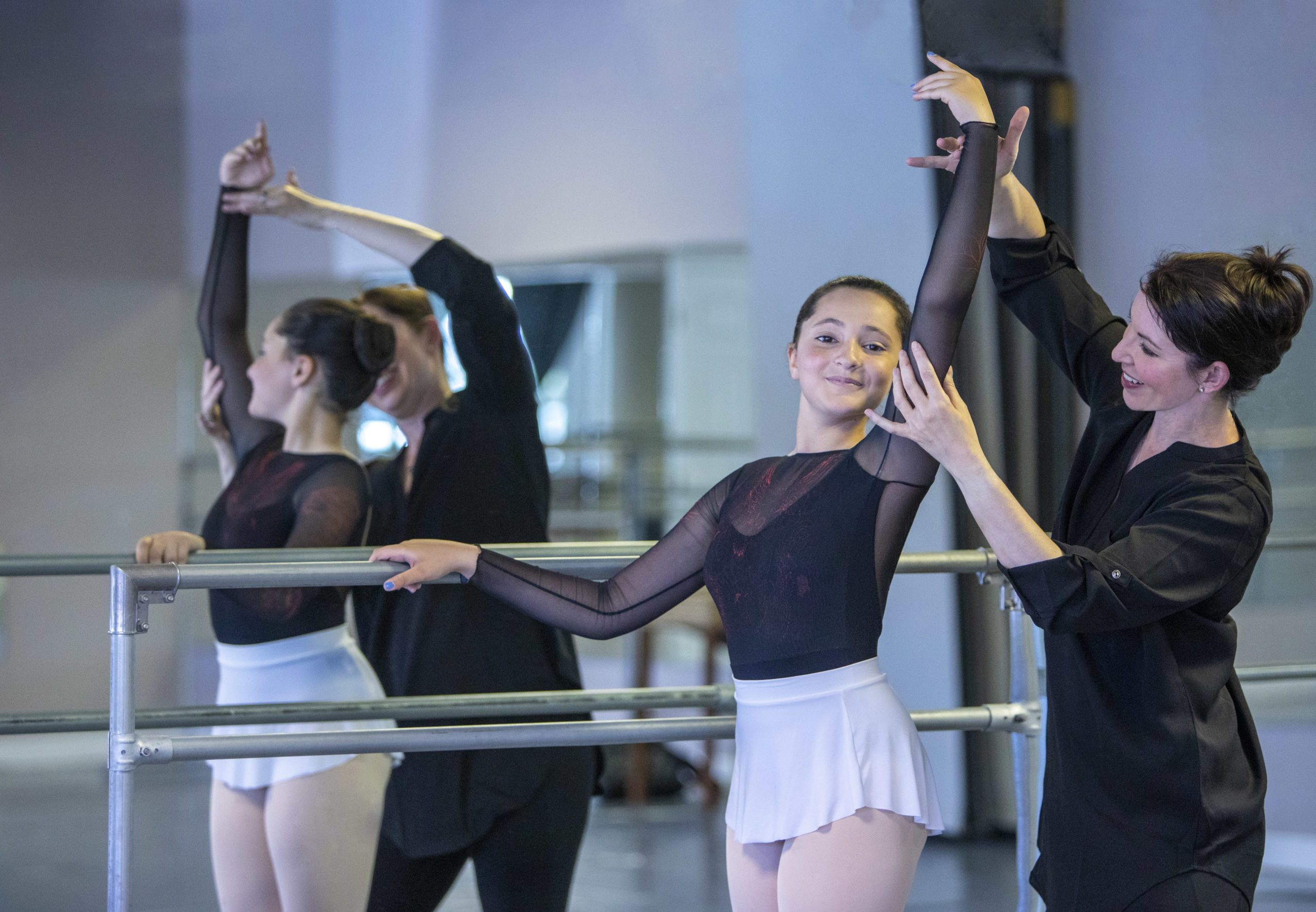 A young ballet student stands at the barre with one arm up in fifth position. She is wearing a black long sleeve leotard and a white ballet skirt. Amy Lowe stands next to her correcting her position, dressed in all black and looking towards her.