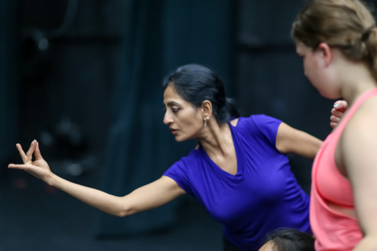 Ananya Chattejea, wearing a bright blueish-purple t shirt, leans over, looking at her hand which is arranged in a intricate gesture. A student in a pink shirt looks on.