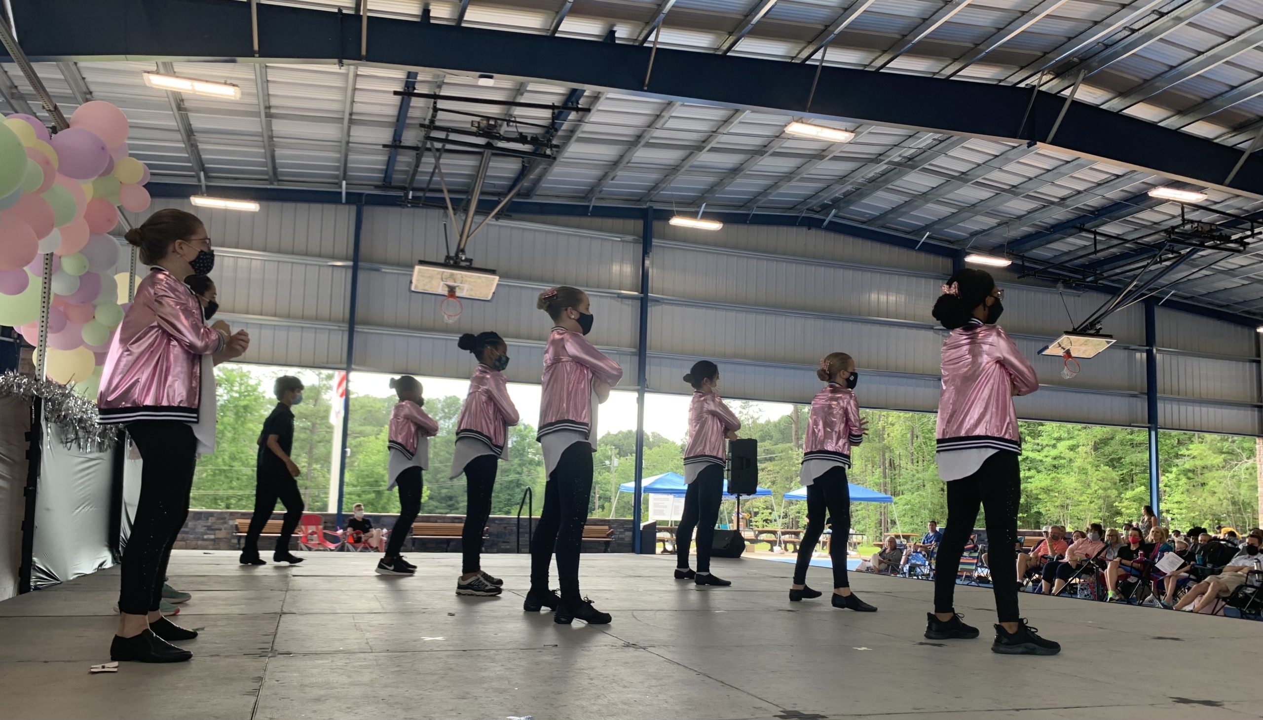 A group of young dancers perform a jazz dance on an outdoor stage, with the audience seated on camping chairs. The young women wear shiny pink jackets, the young man wears all black, and they all wear masks.