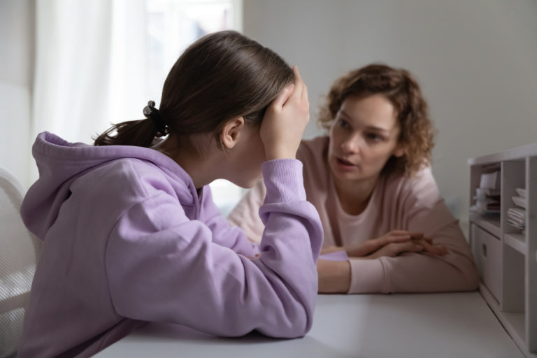 A mother and teenage daughter sit at a table, talking. The daughter looks distressed, putting her hand on her forehead.