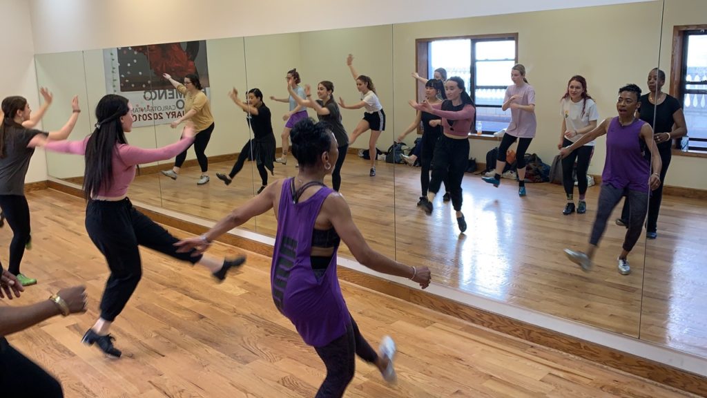 Germaine Goodson, a Black woman in a purple tank top, teaches a tap class. She and the students have one leg extended in front of them. We see them from the back, and their reflections in the mirror.