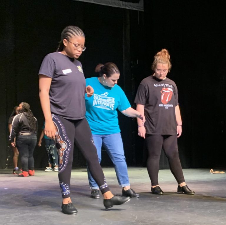 Leia Giddens, a white woman in a blue t-shirt and jeans, teaches two teenage students on a stage. They all wear tap shoes and are looking down at their feet.