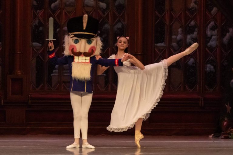 A young dancer plays Clara, in a long white nightgown-like dress, and partners with the Nutcracker, who wears a big Nutcracker head. Clara is in arabesque, holding onto her Nutcracker's arm for support.