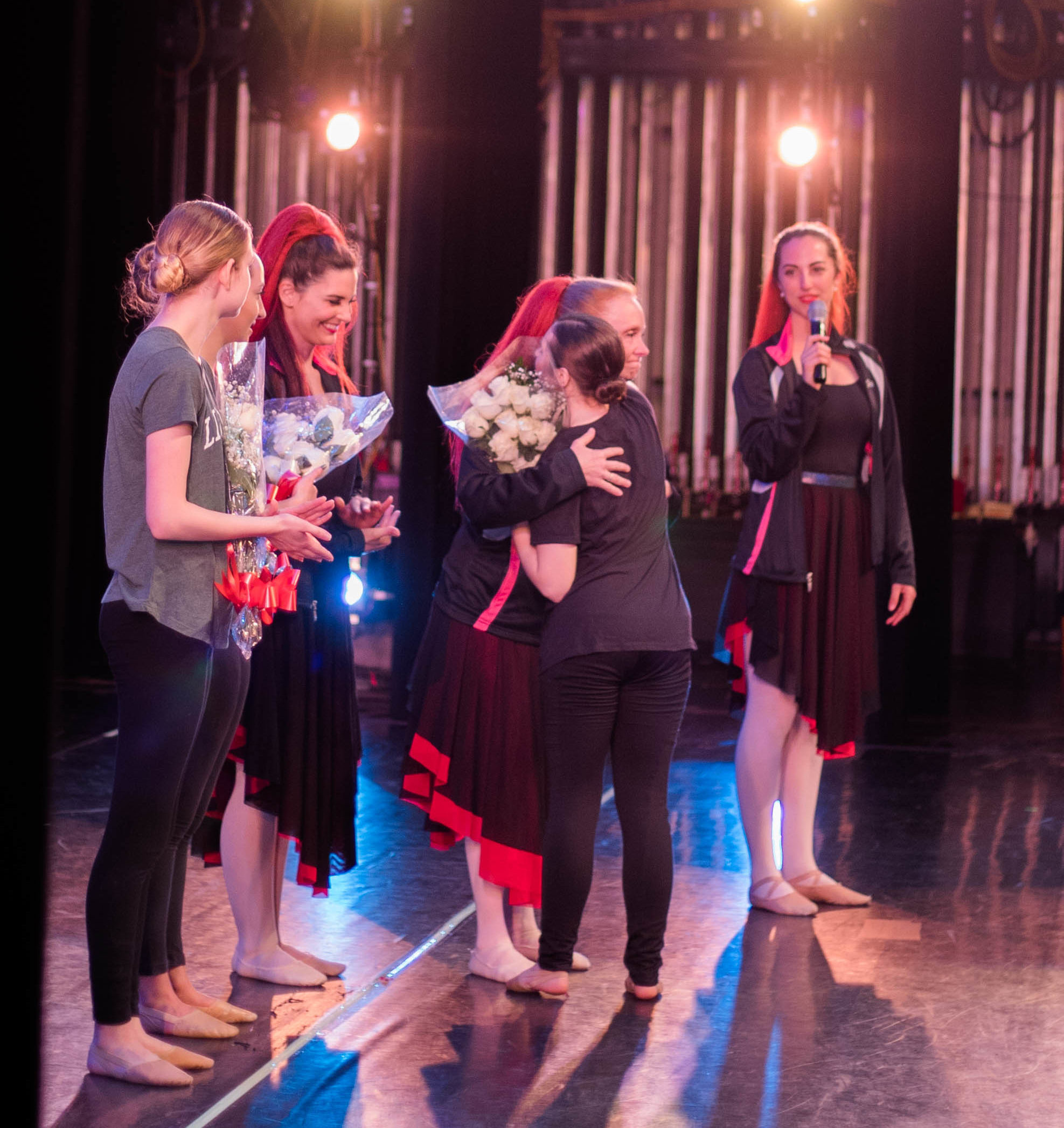 Four dance teachers, in costume from performing, hand out flowers to senior dancers at the end of a recital. They are onstage, with stage lights shining on them, and one student hugs a teacher.