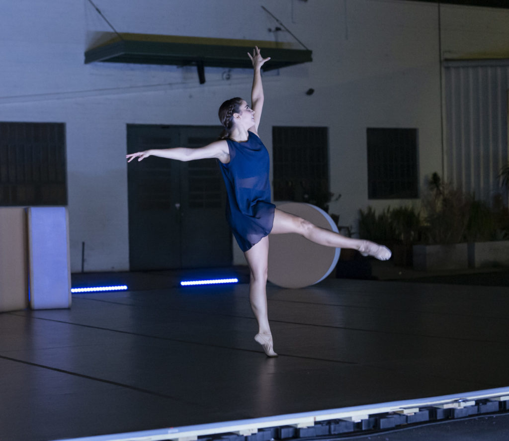 Daisy Jacobson, wearing a short navy dress, performs on a small stage, reaching one arm up and one to the side, and extended one leg forward, while on releve.