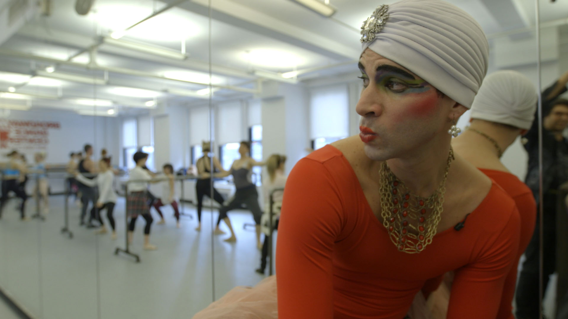 Madame Olga, in a white head wrap, dramatic makeup and an orange leotard, looks over her shoulder at the ballet class happening behind her.