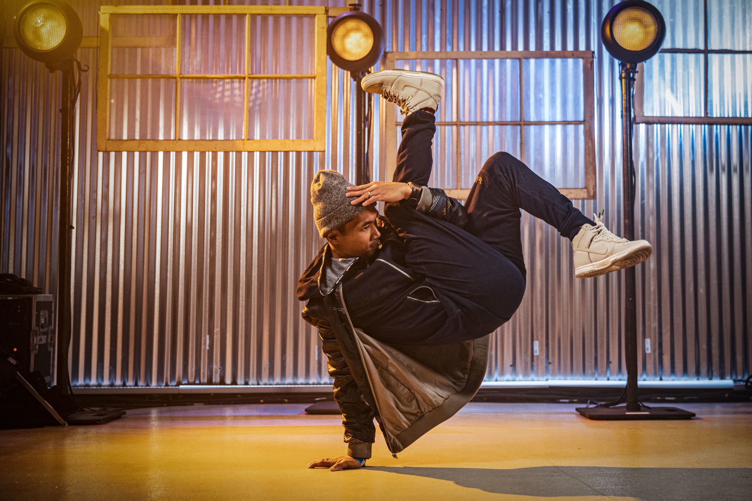 Ronnie Abaldonado balances on one hand, legs kicking up in the air and other hand touching his grey hat. He wears black pants and a green jacket.