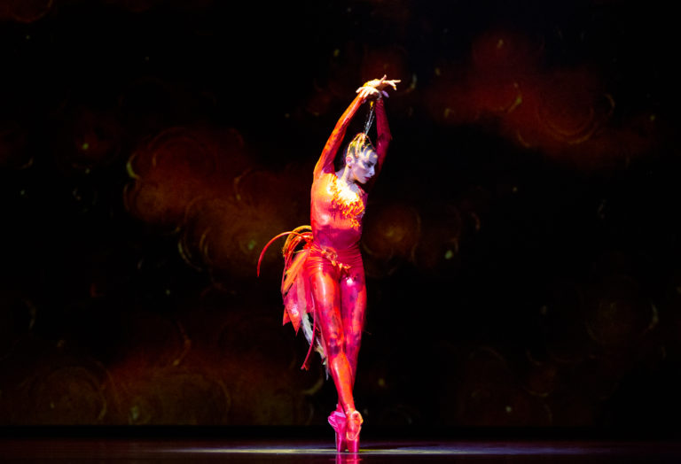 Nathalia Arja, wearing a bright red and orange bodysuit with feathers, leans slightly over, on pointe, arms raised above her head.