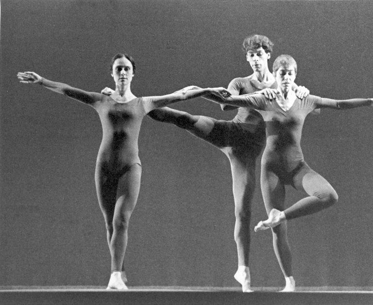 In a black and white archival image, three dancers in identical, long-sleeved unitards intertwine while facing downstage. One woman stands with her feet in a loose fifth, arms extended side. A male dancer balances on relevé with his leg extended side, holding the shoulders of a woman who stands in an overcrossed retiré while holding the hand of the first woman.