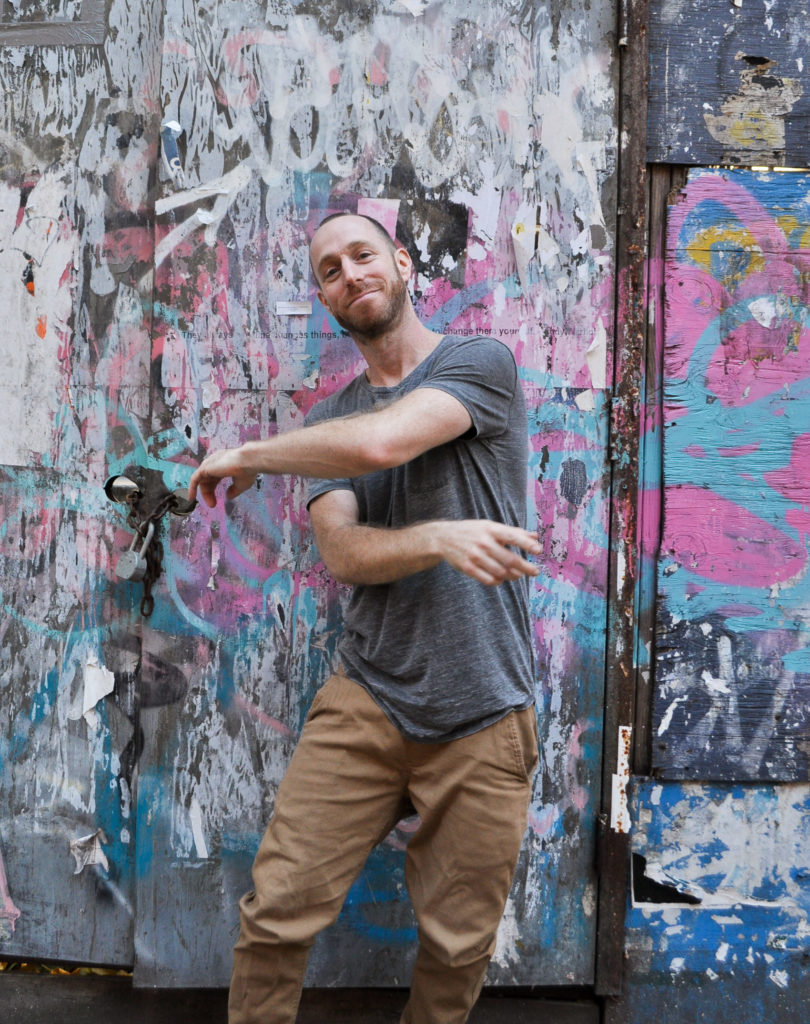 Marc Kimelman, a white man wearing a grey shirt and khaki pants, swings his arms in front of his torso, leaning slightly to the side. He is front of a colorful graffitied wall.