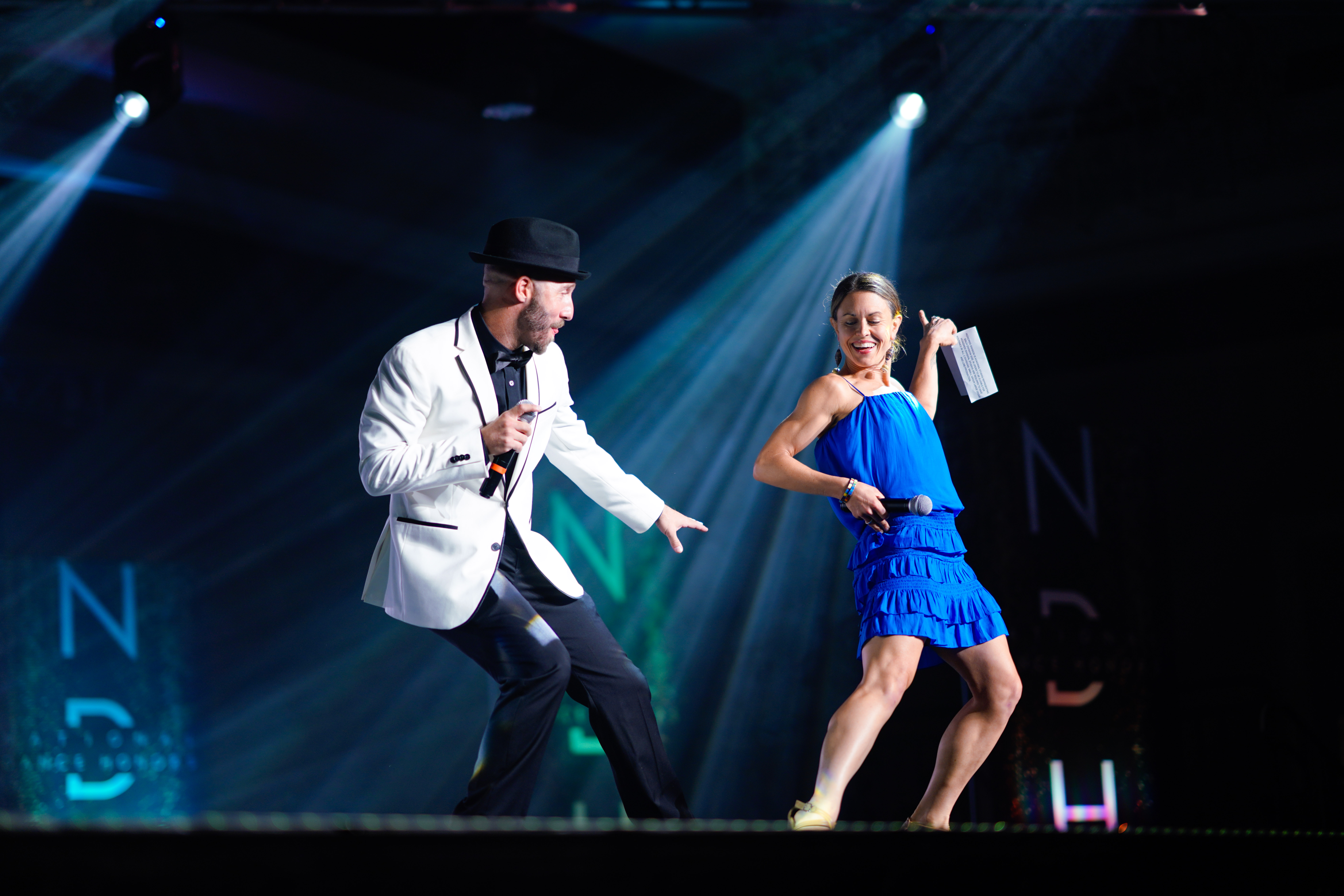 Adrenaline Faculty Members Nick Bass and Caroline Lewis Jones dance together onstage holding microphones. Bass wears a white sport coat and black top hat, and Lewis Jones wears a royal blue dress with a ruffled bottom and heels.