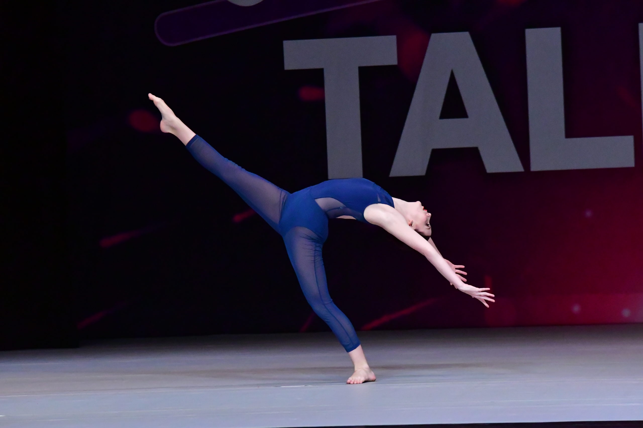 A dancer in a blue leotard and blue tights extends her leg in the air, arching back towards the floor. She is on a competition stage, with the "Applause Talent" backdrop