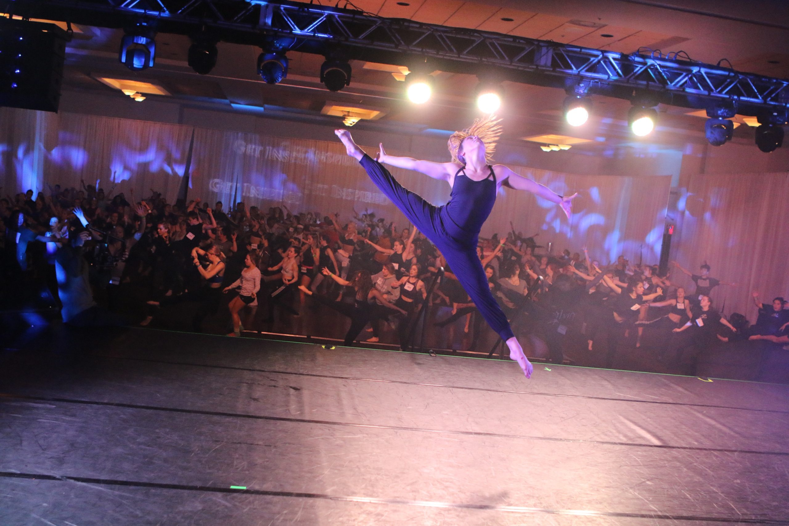 Hailee Payne leaps onstage at a convention, with what looks like hundreds of students watching her from below. She wears all black, and throws her head back as she jumps.