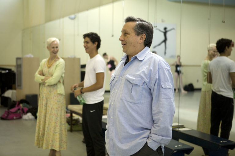 Bo Spassoff wears a loose blue buttom up shirt that is untucked from his dark pants. He stands with his back to a dance studio's mirror looking out into the room with a smile. Behind him is a male student and a woman in a long green dress.
