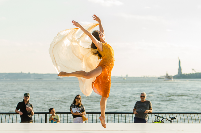 Elizabeth Wright performs on an outdoor stage in front of the river in New York City, the statue of liberty visible in the background. She wears an orange dress with an orange veil attached at her wrists, which flows behind her as she jumps.