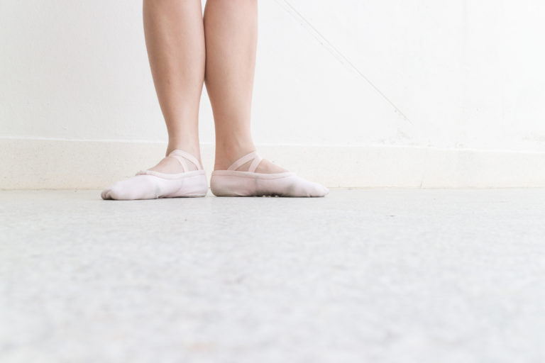 A dancer's feet in first position. Her legs are bare and she wears pink ballet shoes, against a light wall and floor.
