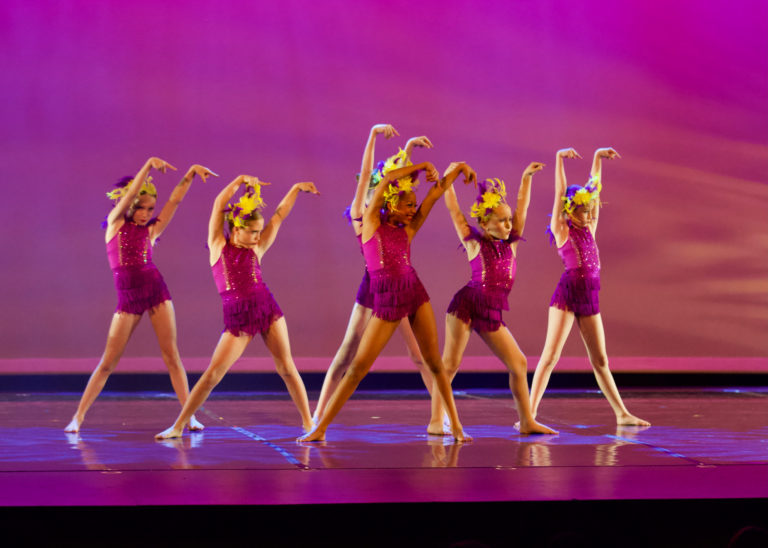 A group of young dancers in pink costumes and yellow headpieces perform against a bright pink backdrop. They raise their arms straight in the air, flexing their wrists.