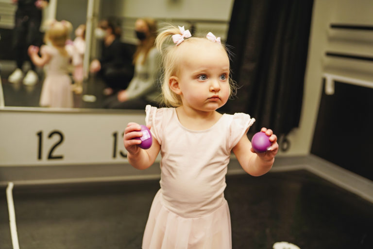 A toddler wearing a pink leotard and pink bows in her hair holds two shakers in her hands.