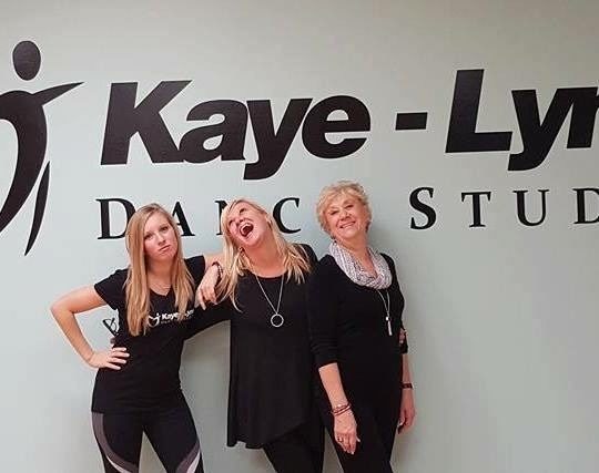 The three women of Kaye-Lynn Dance Studio stand together in front of a wall with the studio's logo. They all wear black and laugh, with arms around each other
