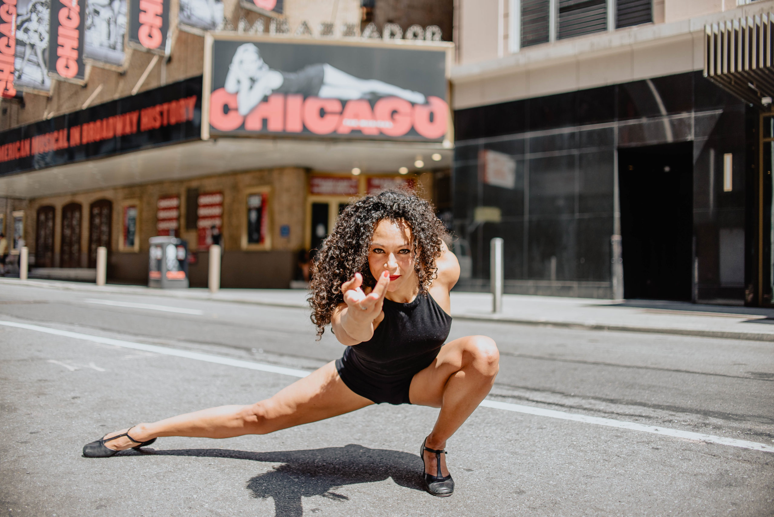 Arian Keddell poses on the street in front of the Chicago theater on Broadway. With one leg extended to the side and one leg bent under her, she hovers close to the ground, reaching one arm forward.