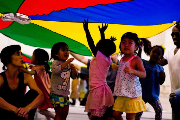 A group of young students play under a colorful parachute, running around, some with their hands in the air