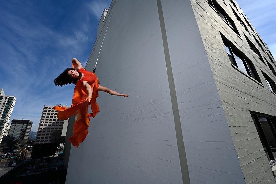 Rose Huey, in a flowing red dress, dances on the side of a white building, suspended from a wire
