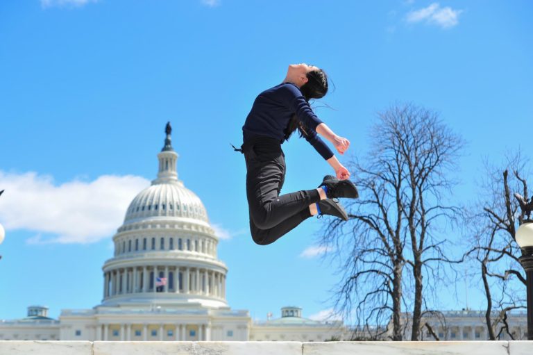 Rosari Sarasvaty jumps in the air in front of the Capital building in Washington, DC. She kicks her legs behind her, bending at the knee, and arches her back. She wears a dark outfit.