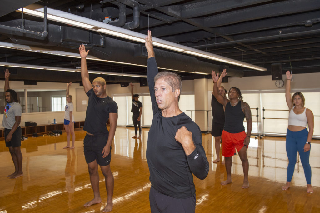Christopher Dolder stands at the front of a dance studio with one arm overhead. He wears long pants and a blac long-sleeve shirt. Behind him is a group of student athletes who are taking his modern dance class, mimicking the one-arm-overhead pose. The group is a mix of men and women and all wear various workout clothes.