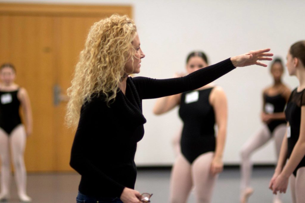 Meg Paul stands in profile with her left arm lifted to point at something off camera. Her wavy blonde hair is down around her shoulders and she wears a black long-sleeved shirt. Behind her is a student in a black leotard and pink tights.