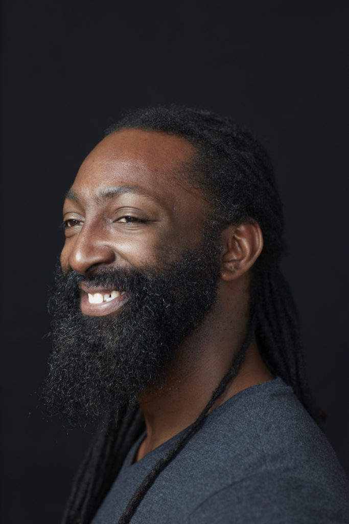Antoine Hunter: A Native American African-American person with dark chocolate skin from his mother. He has almond shaped eyes with long lashes. He has long black dreadlocks tied in a low braid and a full beard. Antoine is wearing a dark v-neck shirt and smiling.