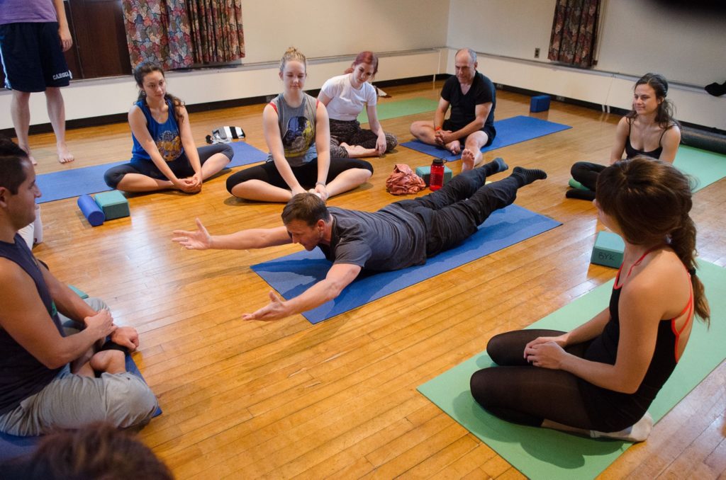 Robert Cook lays face down on a yoga mat in the middle of a dance studio, holding a superman post. He wears long dark pants and a gray t-shirt. Around him, college students dressed in various workout regalia sit on their own yoga mats and watch Cook.