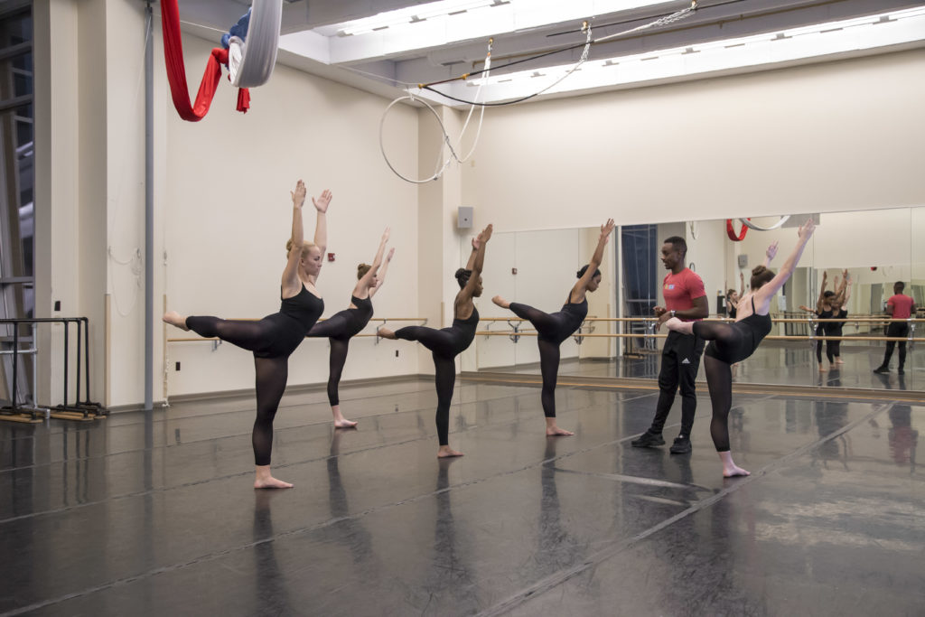 Kirven Douthit-Boyd stands in the middle of a dance studio surrounded by six female students. The students wear black leotards with black tights and stand barefooted in a modern-dance arabesque. Kirven wears black pants and a red shirt.