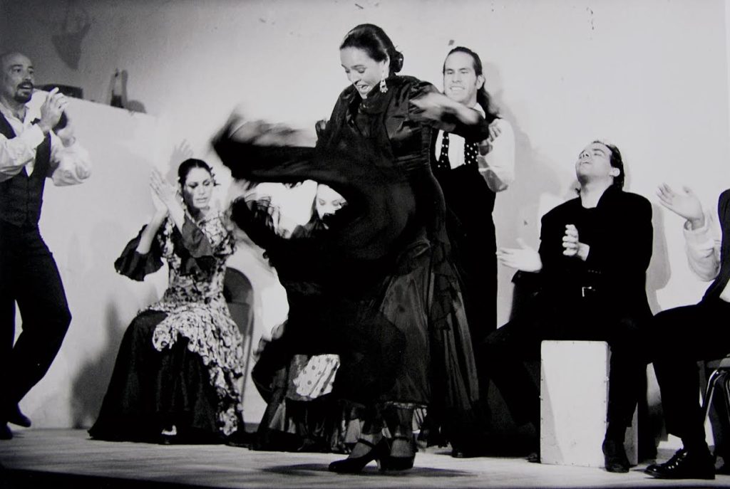 In a black and white image, a young Eva Encinias grins, eyes lowered as she whirls her skirt onstage, feet close together. Arrayed around her are other singers and dancers clapping along to her performance.