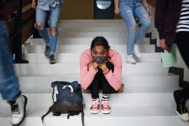 A young African American student sits on the steps of a school hallway. She wears red sneakers and patterned mask.
