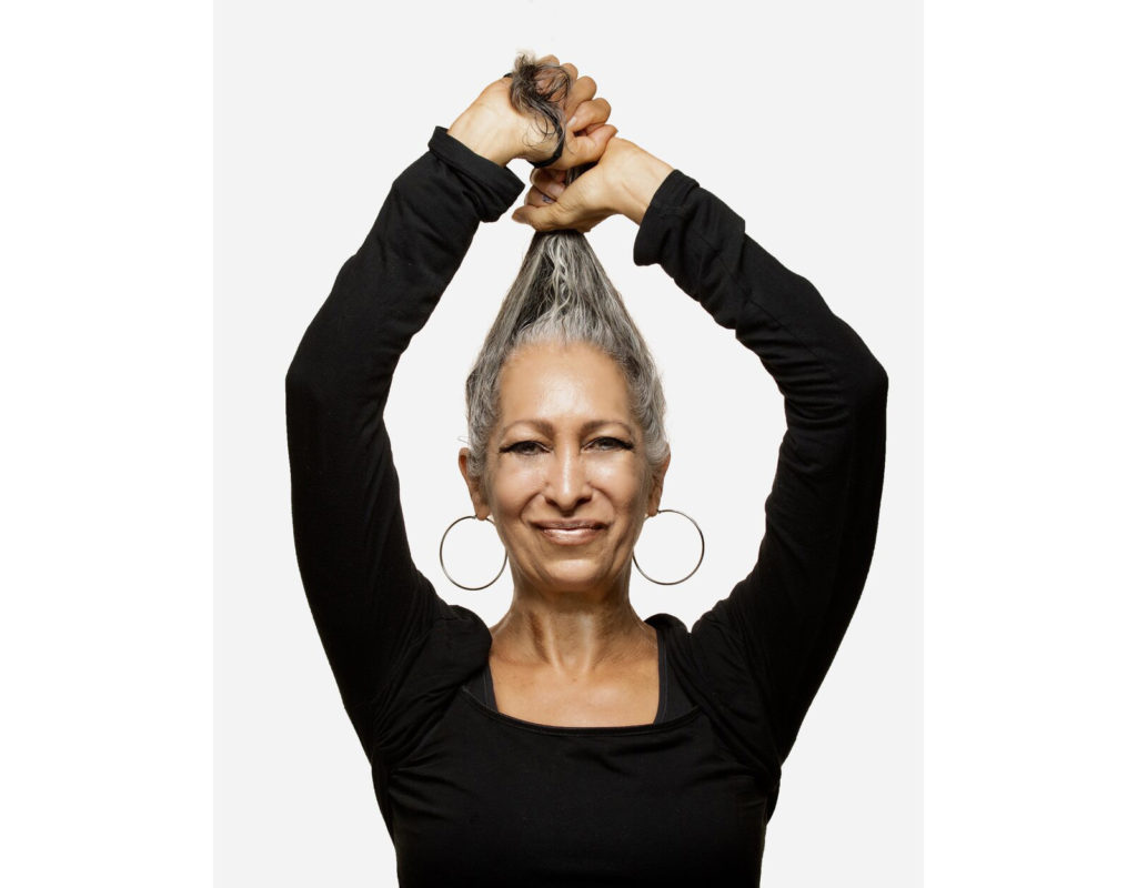 Cynthia King, wearing a long-sleeved black shirt and large earrings. She pulls her salt and pepper hair up above her head and smirks.