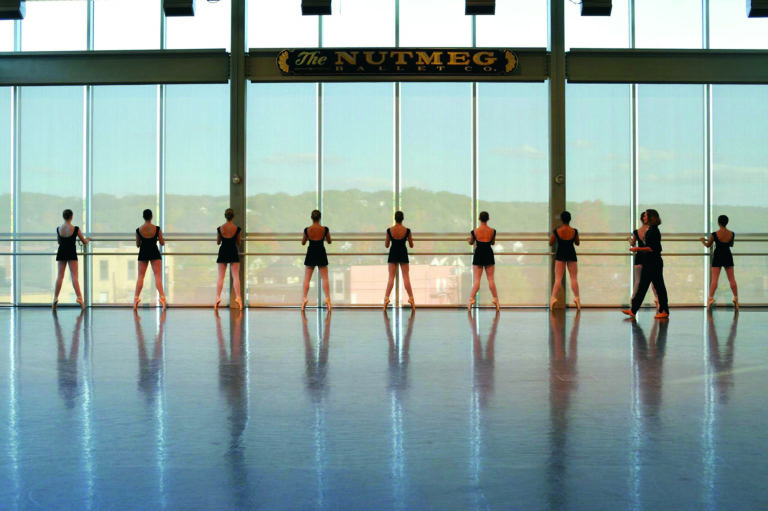 A line of ballet students facing the barre in a ballet studio. The barre is attached to a large floor-to-ceiling window, and the light pours in over the students.Victoria Mazzarelli walks behind them on the right.