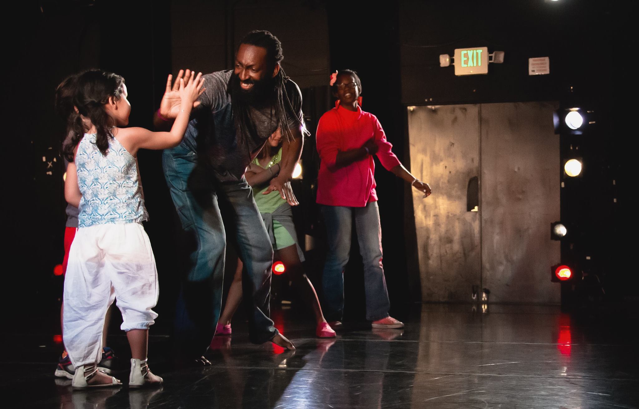 Antoine Hunter, a Native American African-American person with dark chocolate skin from his mother, almond shaped eyes with long lashes, and black dreadlocks and a full beard, leans forward to high five a young dancer.