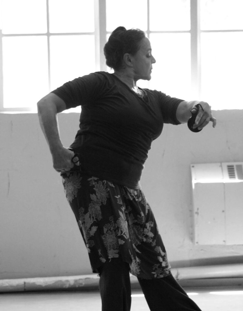 In a black and white image, Eva Encinias is show in profile in a sunlit studio. She pulls her right hand back to her hip, elbow raised, as she looks over her left arm, curving in front of her chest. She pulls her weight onto her back foot, even as her posture pulls her forward.