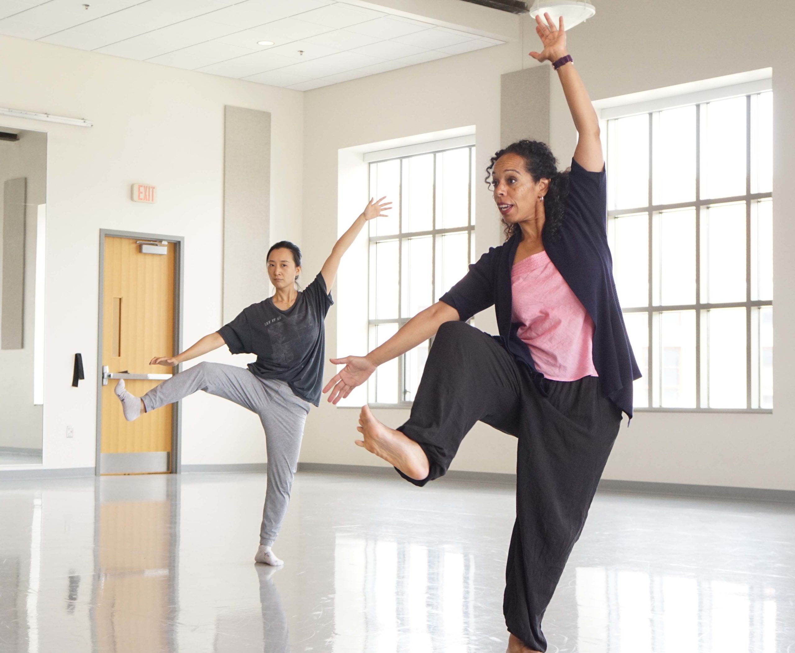 In a sunlit studio, Dr. Nyama McCarthy-Brown, dressed in loose black sweats and a jacket over a pink shirt, dark curls loosely pulled back from her face, demonstrates for a college-aged dancer. She moves through plié, her right knee pulling to her chest with a flexed foot. Her right arm is open at a downward diagonal to one side, while her left reaches with an open palm over head.