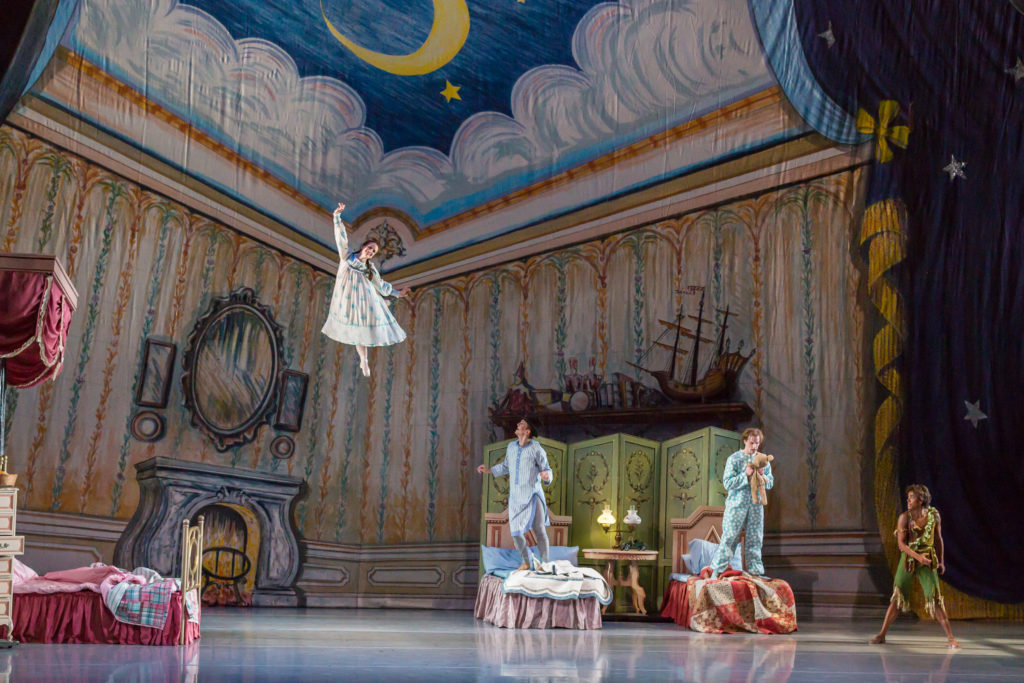 A woman dressed as Wendy in Peter Pan flies above the stage while Michael, John and Peter Pan look up at her from the floor. The set is dressed as a bedroom, with three children’s beds and a big fireplace