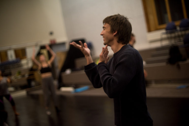 Scotty Hardwig wears a dark longsleeve shirt and stands in the middle of a bright dance studio. He smiles while he gestures one hand to his chin and the other out in front of his mouth. Behind him you can see dancers working out of focus with the camera.