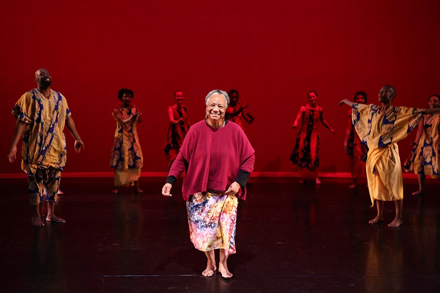 A picture of Kariamu Welsh facing front walking on stage with dancers around her—all against a red background