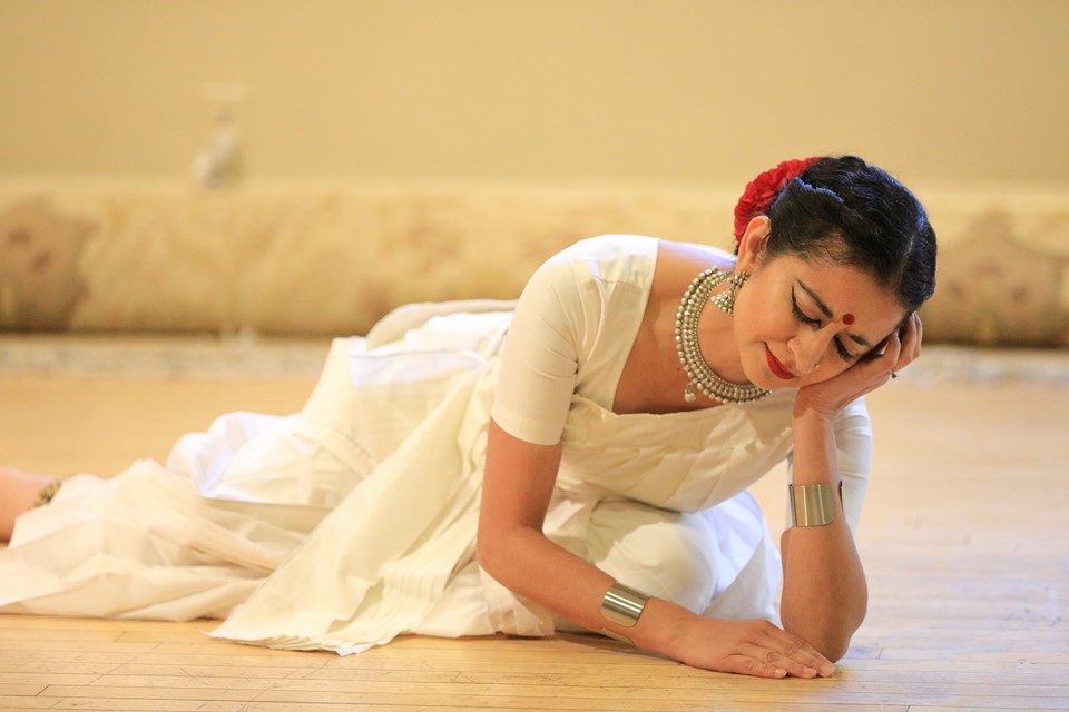 Image of an indian dancer in a white saree with a silver necklace and red flower in her hair, sitting on the ground with one hand to her cheek and the other resting on the floor