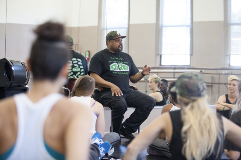 Image of acclaimed choreographer Rennie Harris sitting on a chair and instructing his dance students in a studio