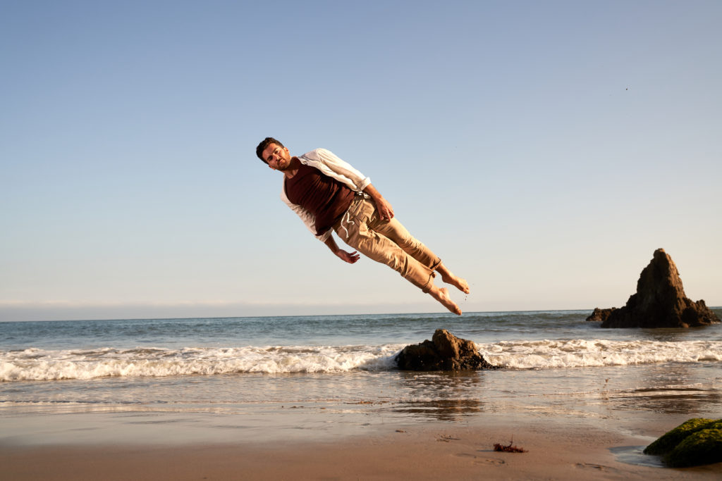 A male dancer elevated off the ground and leaning sideways to the left in an empty beach