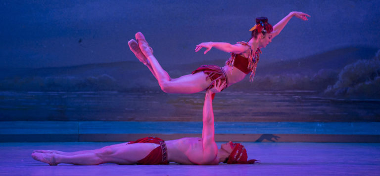 A male ballet dancer lying down and lifting a female ballet dancer—both on stage against a blue background