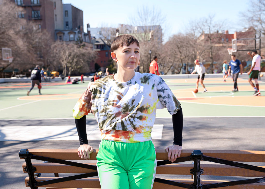 Katy Pyle, wearing a tie dye shirt and bright green shorts, leans against a park bench next to a basketball court.