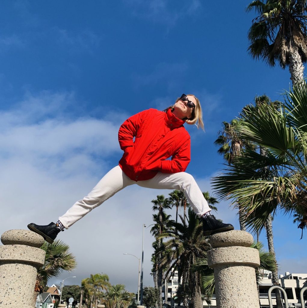 Kelli Erdmann, a young white woman, wears a red sweatshirt and white pants. She stands on two pillars, one foot on each. There are palm trees and a blue sky in the background