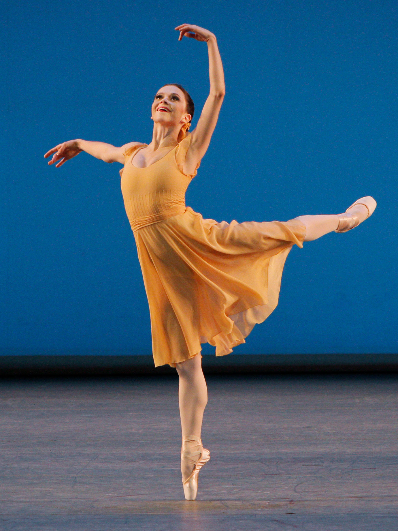 NYCB principal dancer Megan Fairchild in Dances at a Gathering wearing an orange dress against a blue background. She holds an attitude derriere with arms in fourth